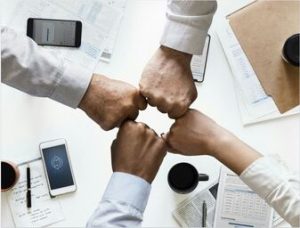 four hands in fists bumping together to form a square over a meeting table with phones, papers, note pads, and coffee cups